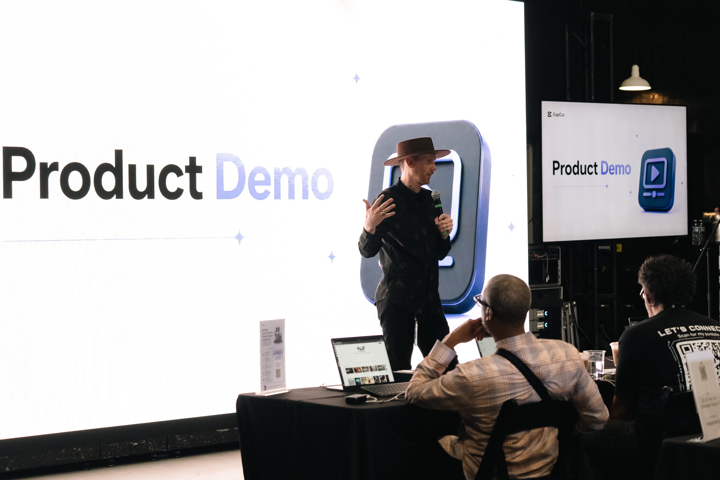 A presenter in a wide-brimmed hat demos on a mic before a huge white "Product Demo" screen, attendees seated below.