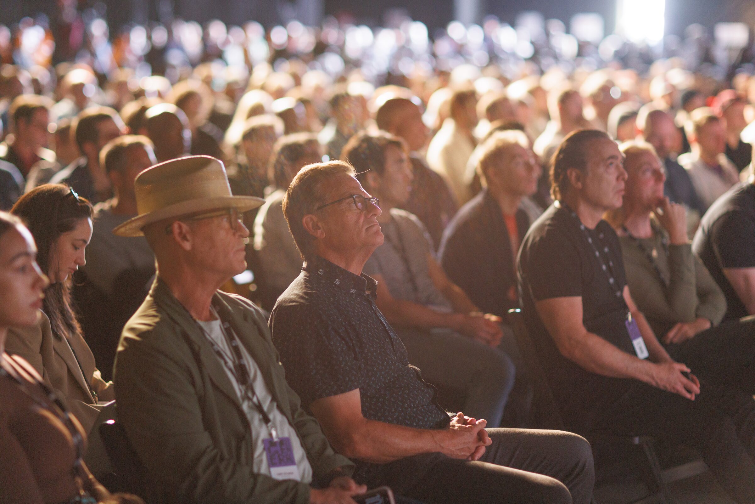 Close-up of seated audience members, one in a wide-brimmed hat, listening intently in a crowded hall.
