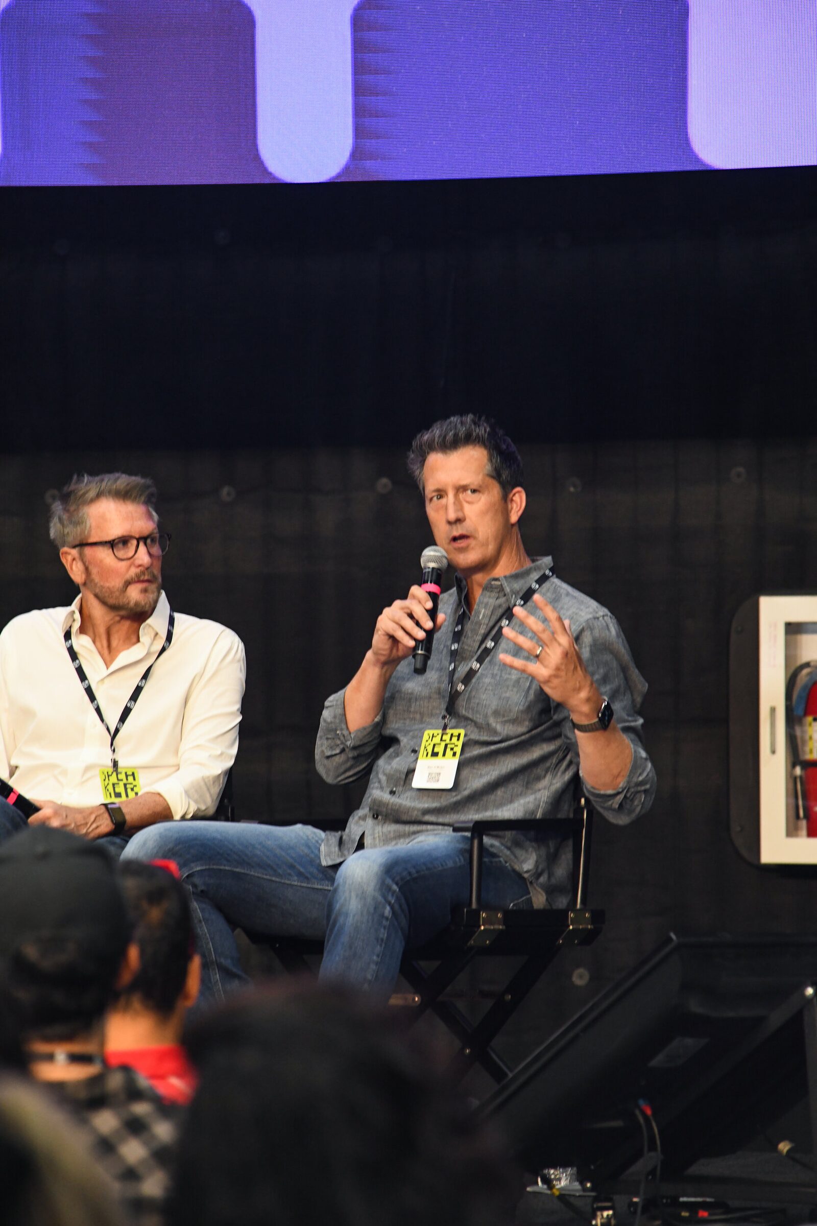 Dan O'Brien, in a denim shirt, talks into a mic in a director's chair, a panelist in white seated beside him.