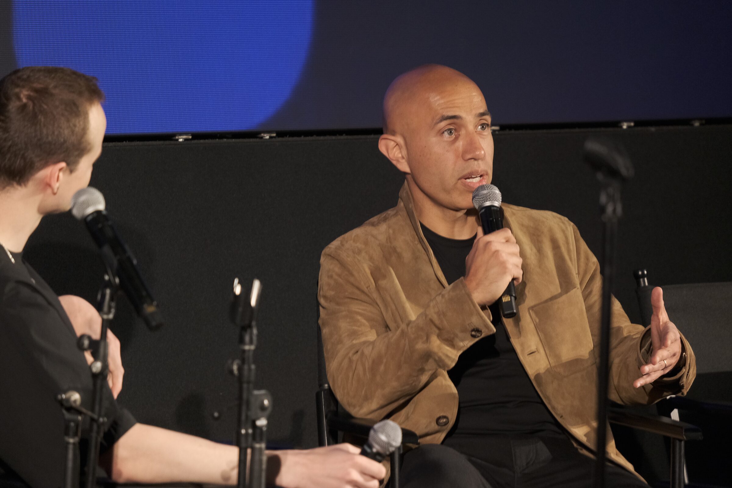 Soleio Cuervo, in a tan suede jacket, speaks into a mic beside a fellow panelist on a dark stage.