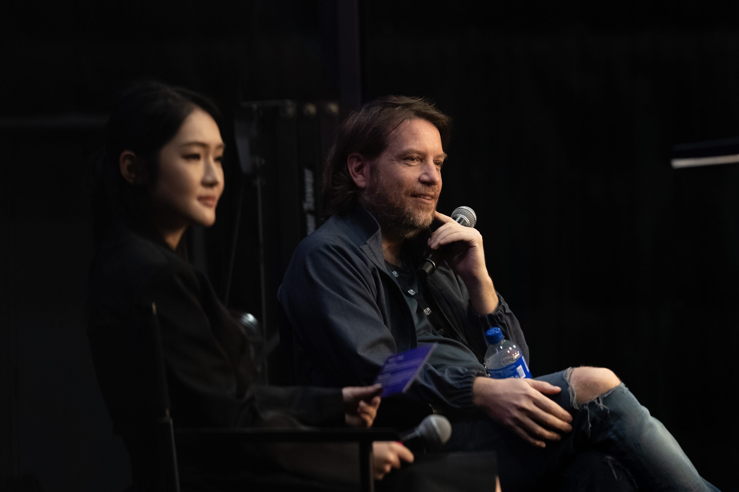 Gareth Edwards smiles and Zoey Zhao listens, both holding mics, as they take a question from the dark audience.