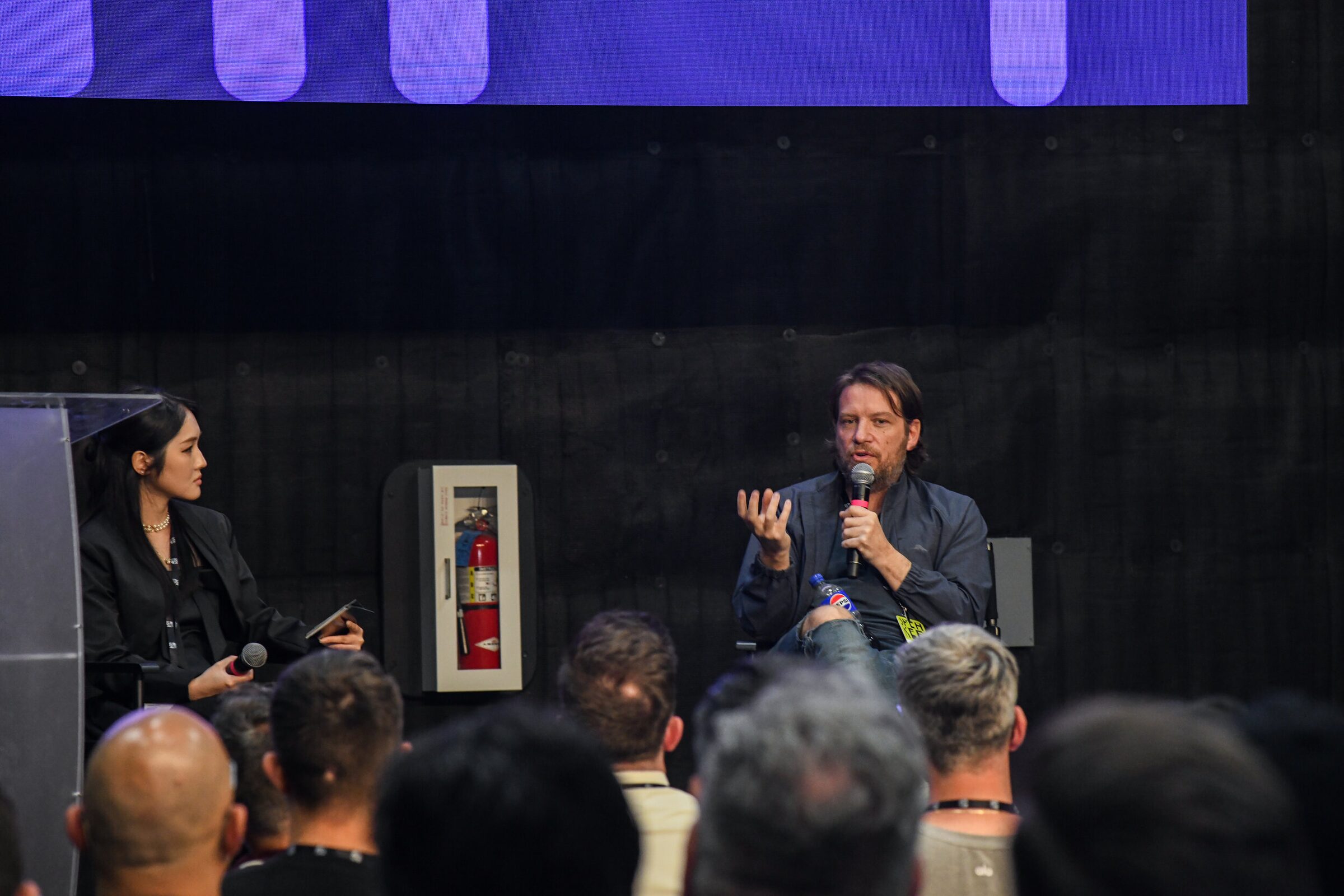 Gareth Edwards gestures with a mic while Zoey Zhao, in black, sits beside him on a dark stage before the crowd.
