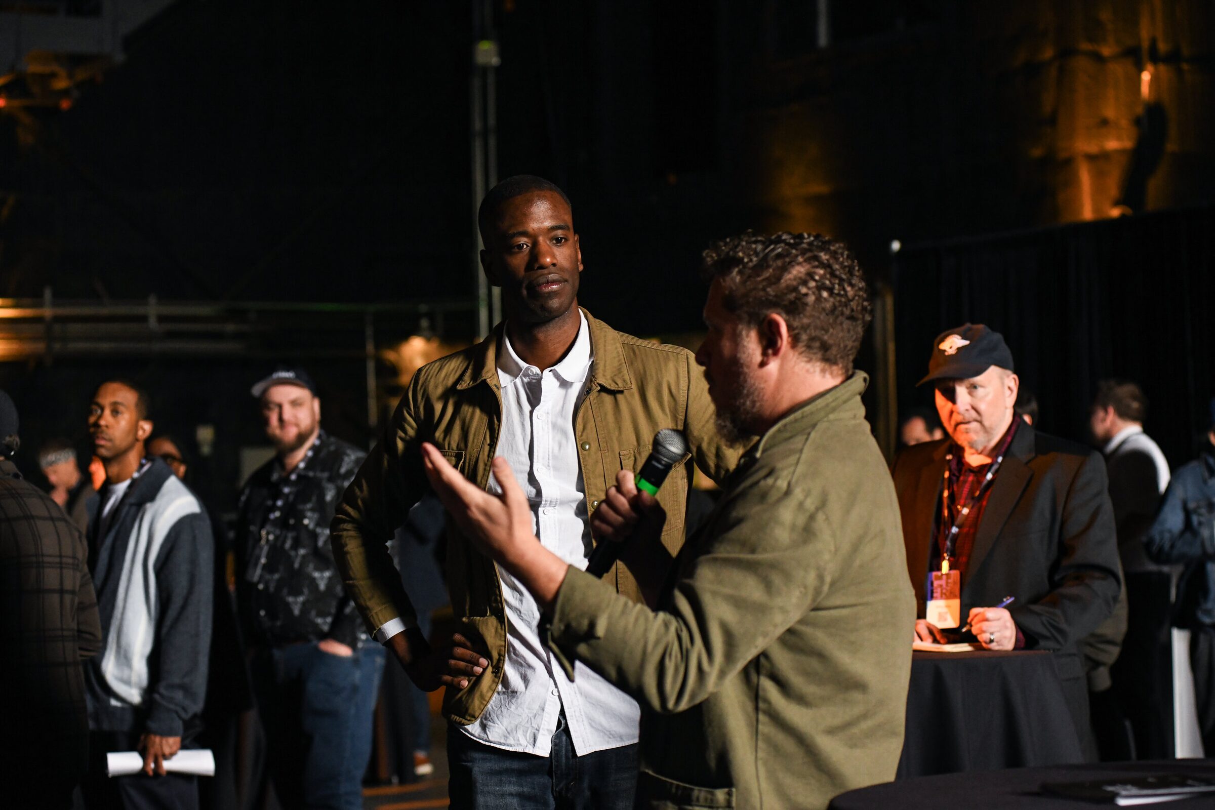 Gaz Alazraki gestures as he asks a question into a mic, Maxwell Kangkolo holding it for him and watching.