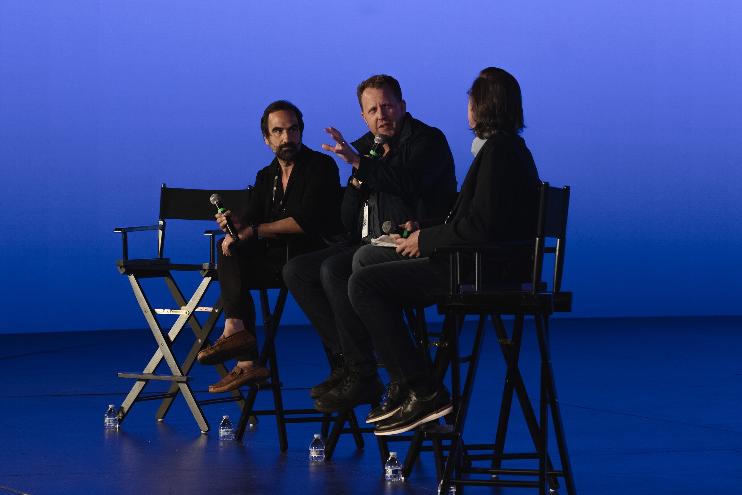 Jon Erwin gestures mid-answer between two fellow panelists in director's chairs against a deep blue backdrop.