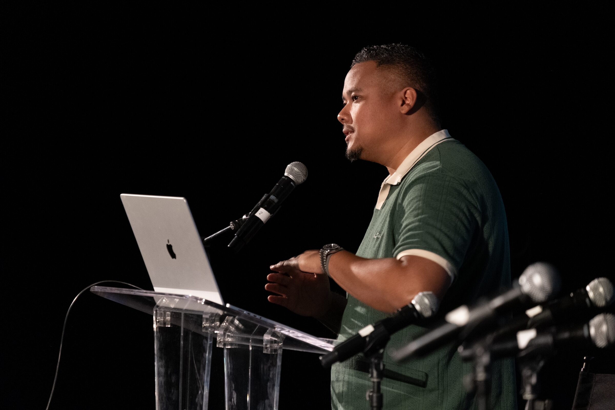 Dave Clark speaks at a clear lectern with a laptop, a cluster of microphones in front of him.