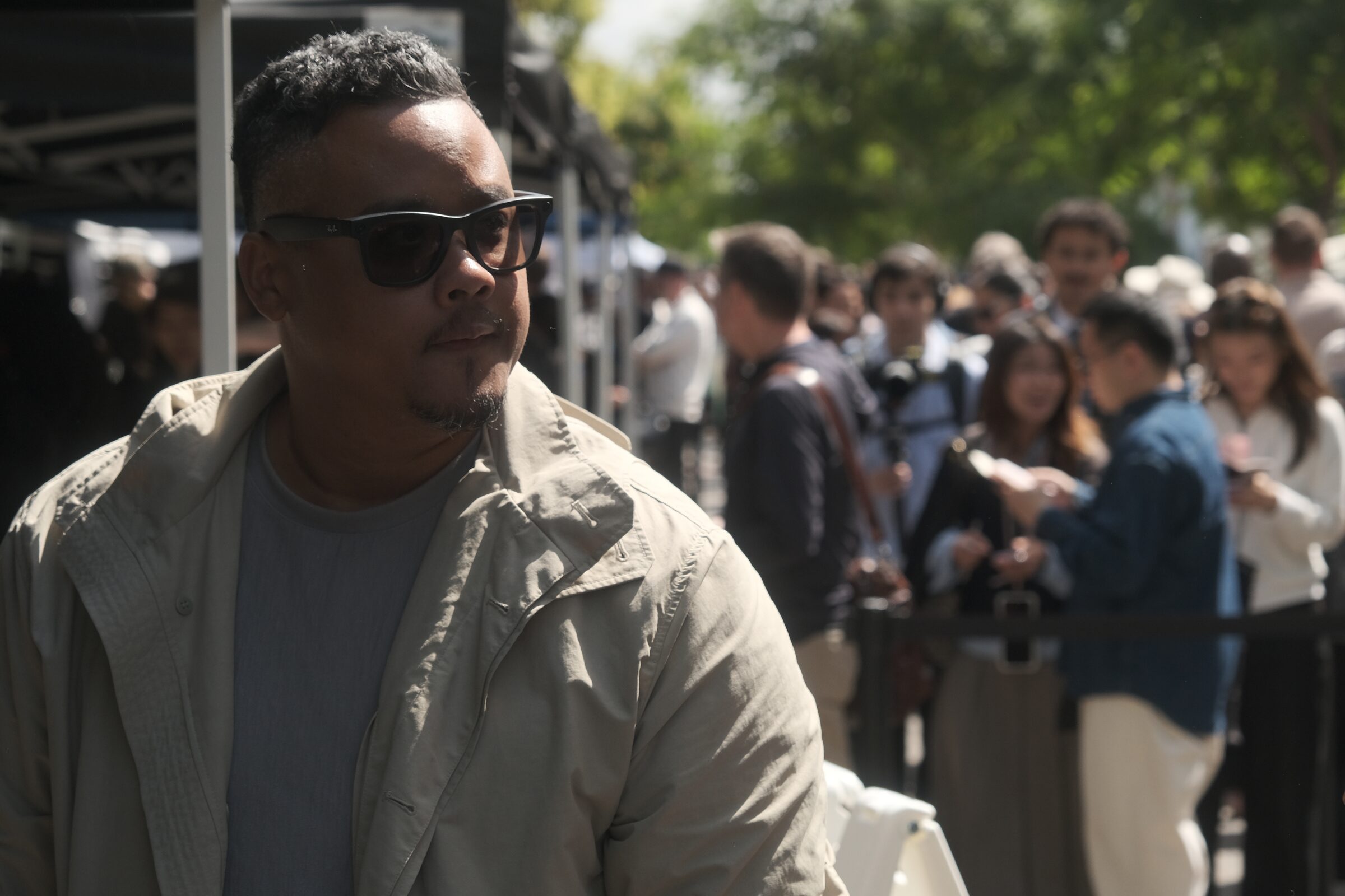 Dave Clark, in sunglasses and a light jacket, looks out over a busy line of attendees outdoors.
