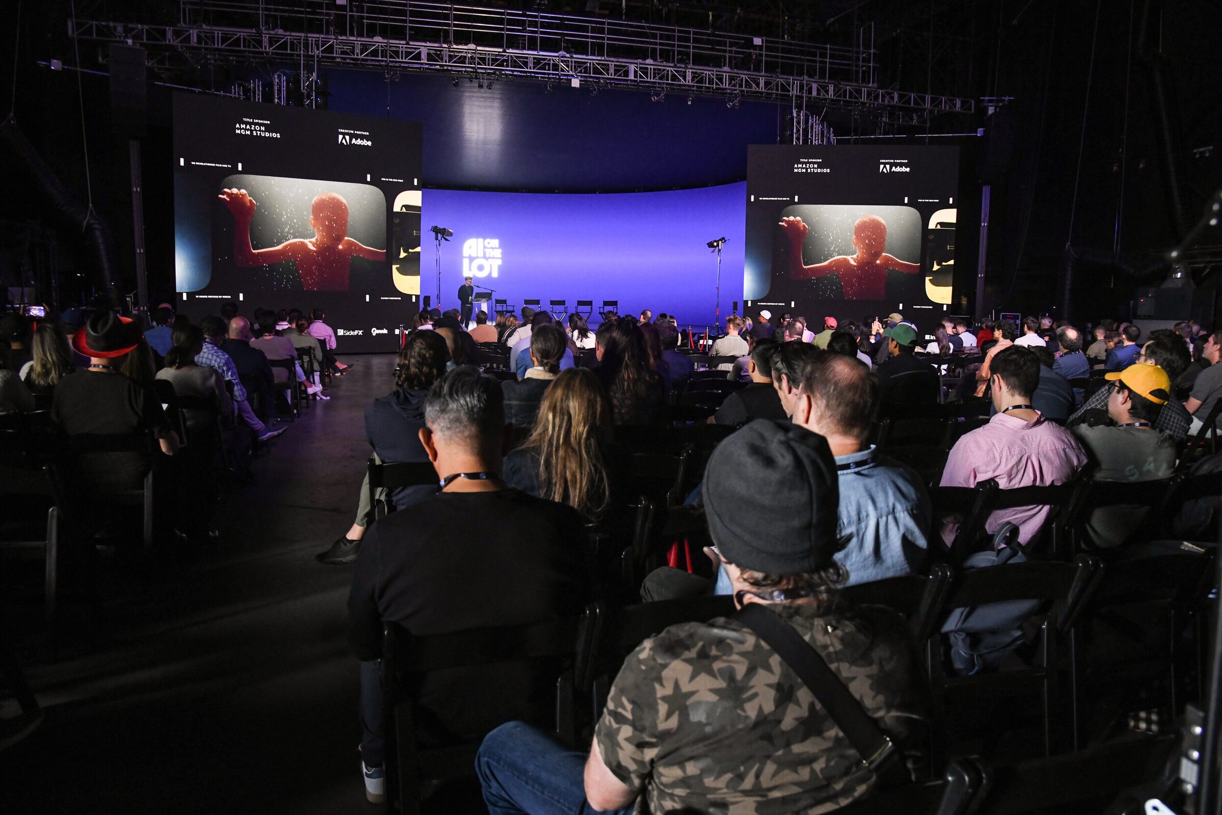 A packed audience faces the curved blue main stage, its twin side screens showing a red-lit film still.