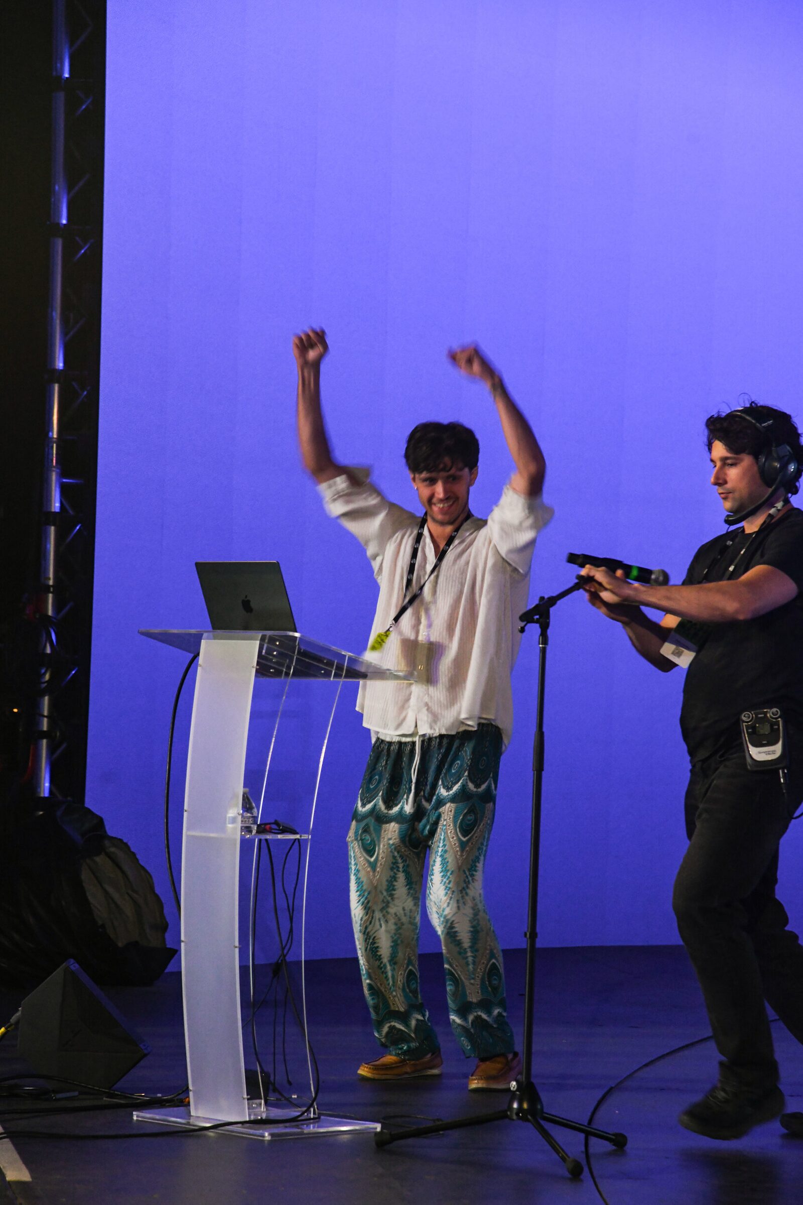 Cameron Kostopoulos throws both arms in the air at the lectern as a stagehand with a headset adjusts the mic.