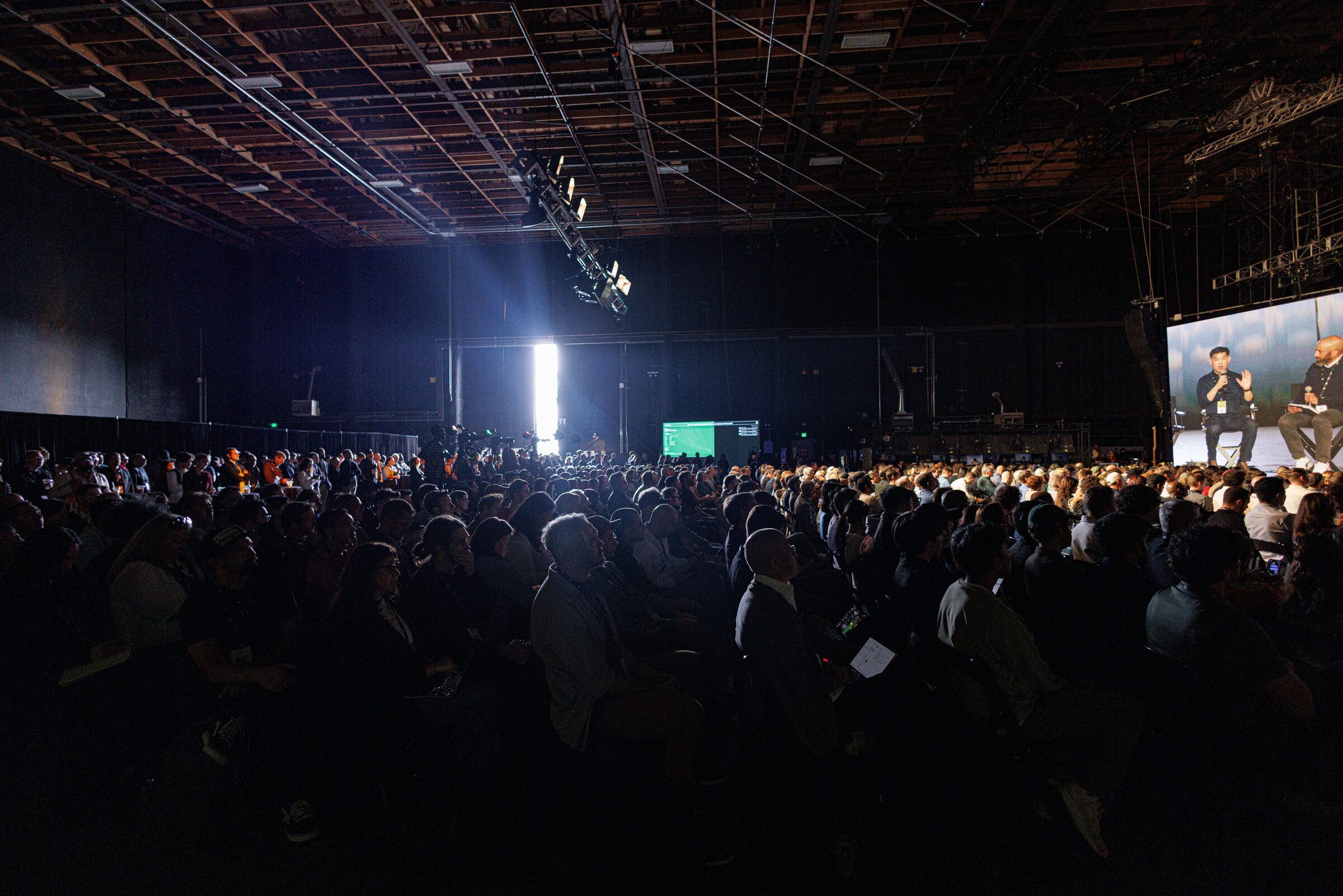 A packed, dark Stage 15 audience watches Albert Cheng and a fellow speaker, shown on a big side screen.