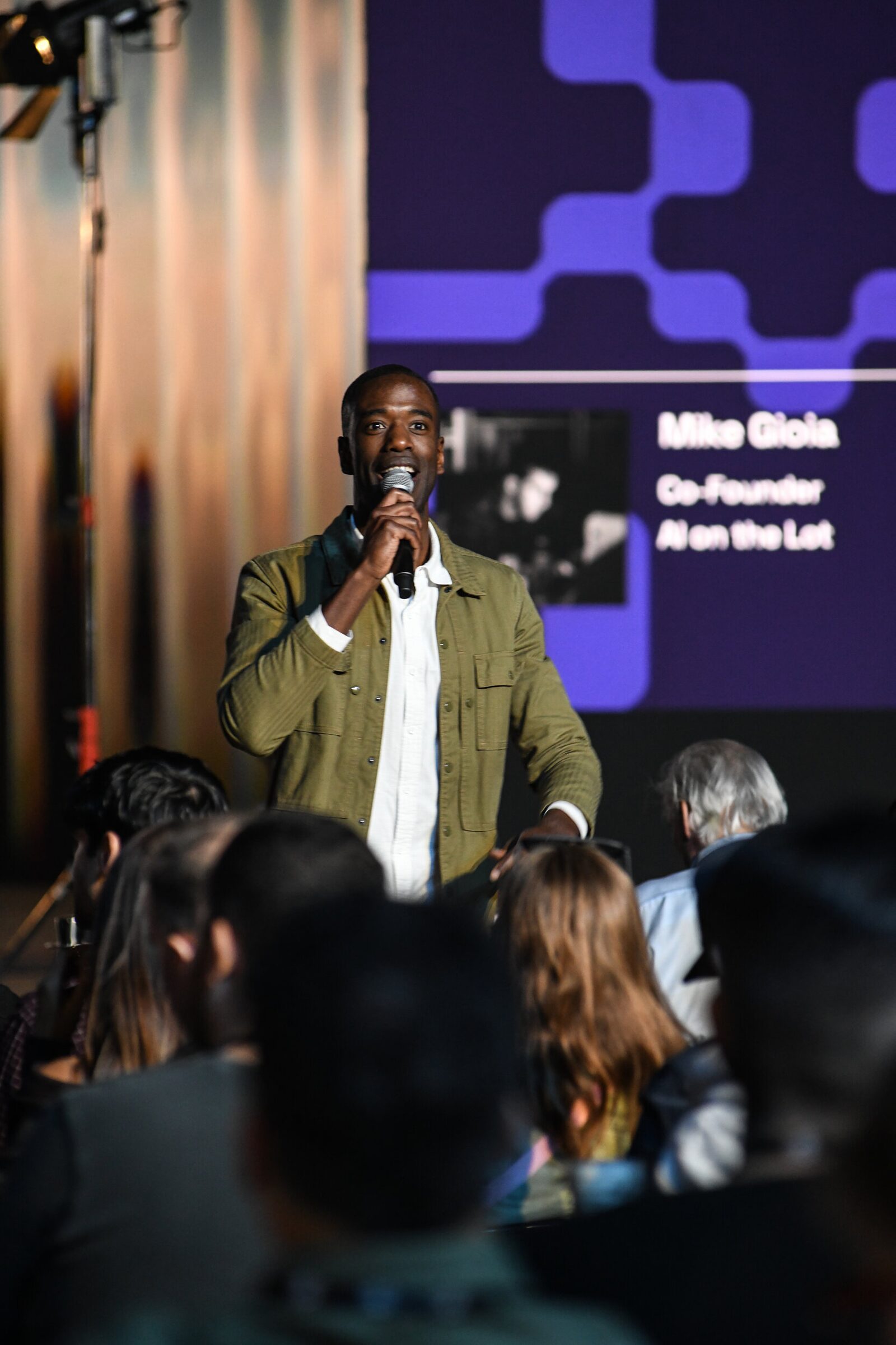Maxwell Kangkolo, in an olive jacket, speaks into a mic in the middle of the seated audience.