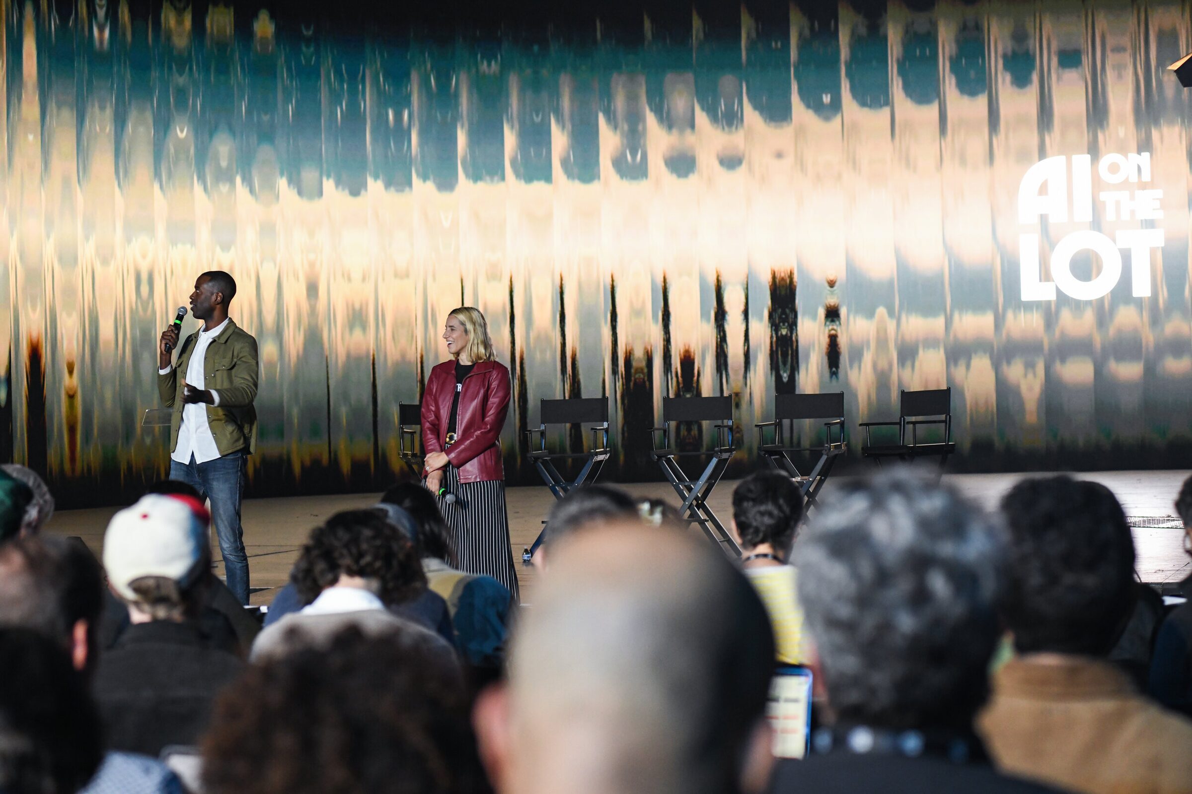 Maxwell Kangkolo and Verena Puhm with mics on the stage, seen past rows of audience heads.