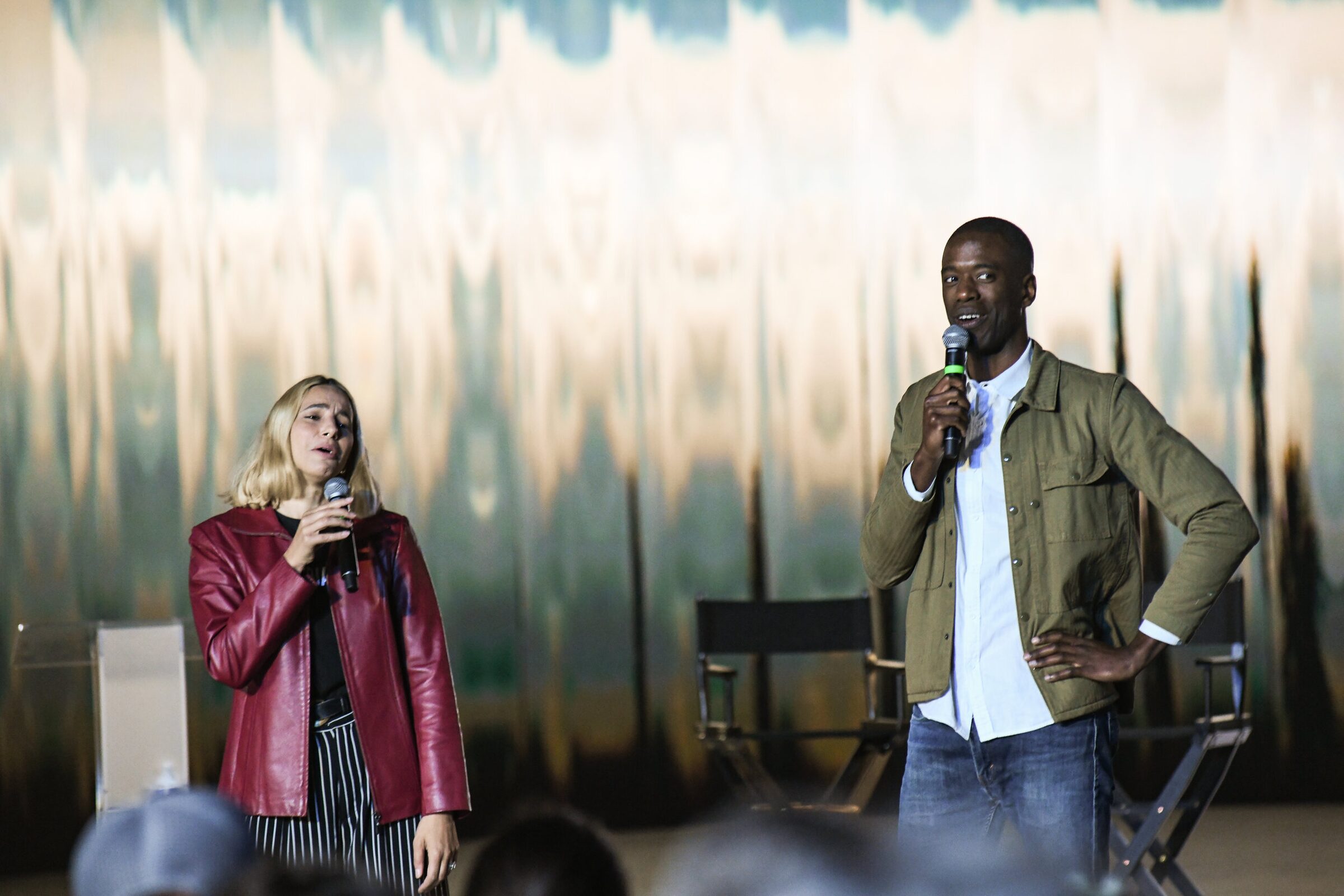 Verena Puhm and Maxwell Kangkolo talk into mics on stage, he with a hand on his hip, in front of a shimmering wall.