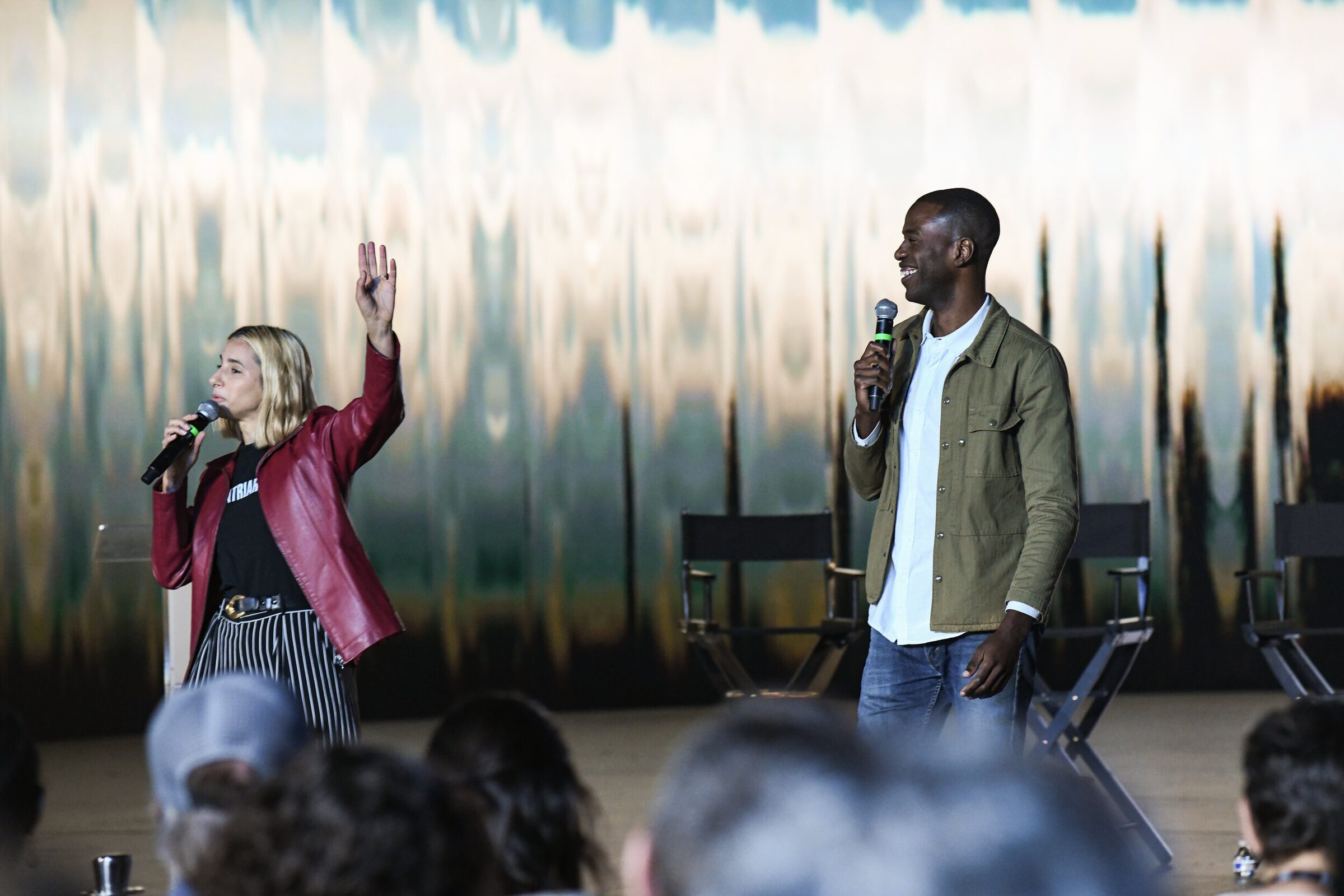 Verena Puhm, in a red leather jacket, raises her hand to the crowd as Maxwell Kangkolo looks on, mic in hand.