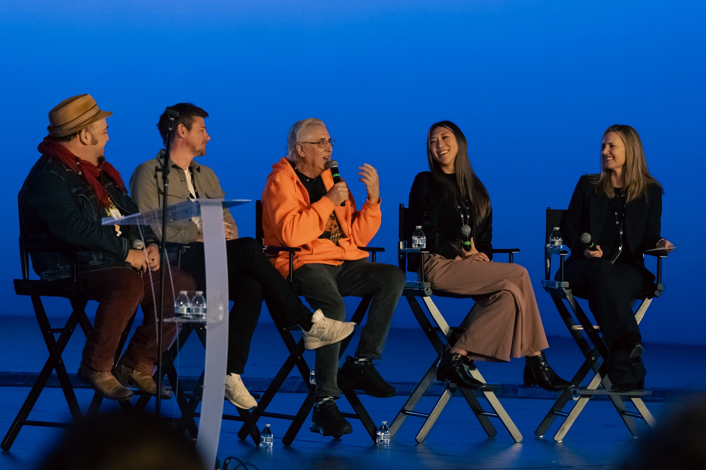 Jessica Fan smiles at the panelist in an orange hoodie as he talks, the five seated across a blue stage.