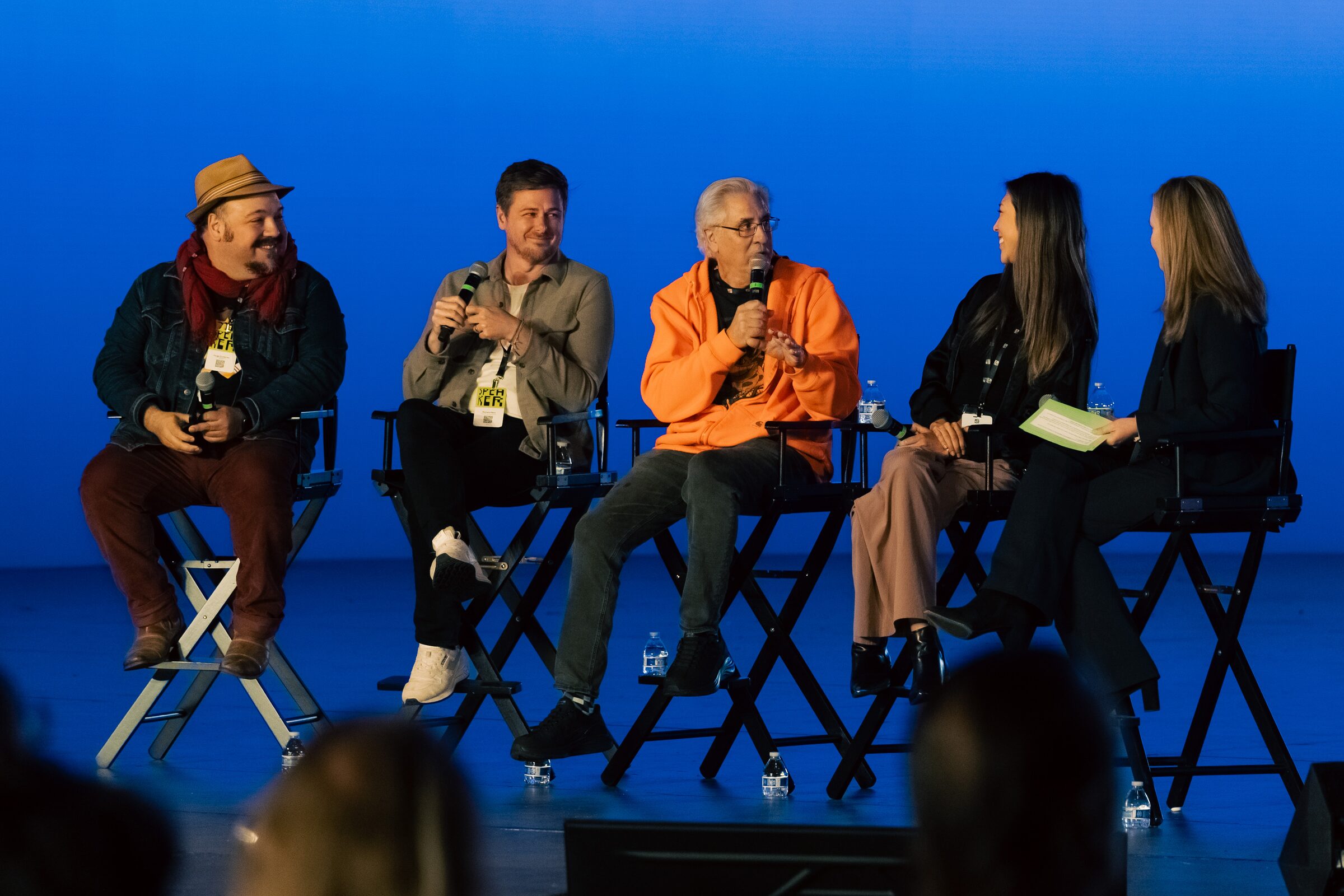 Five panelists in director's chairs on a blue stage, one in an orange hoodie speaking, one in a straw hat.