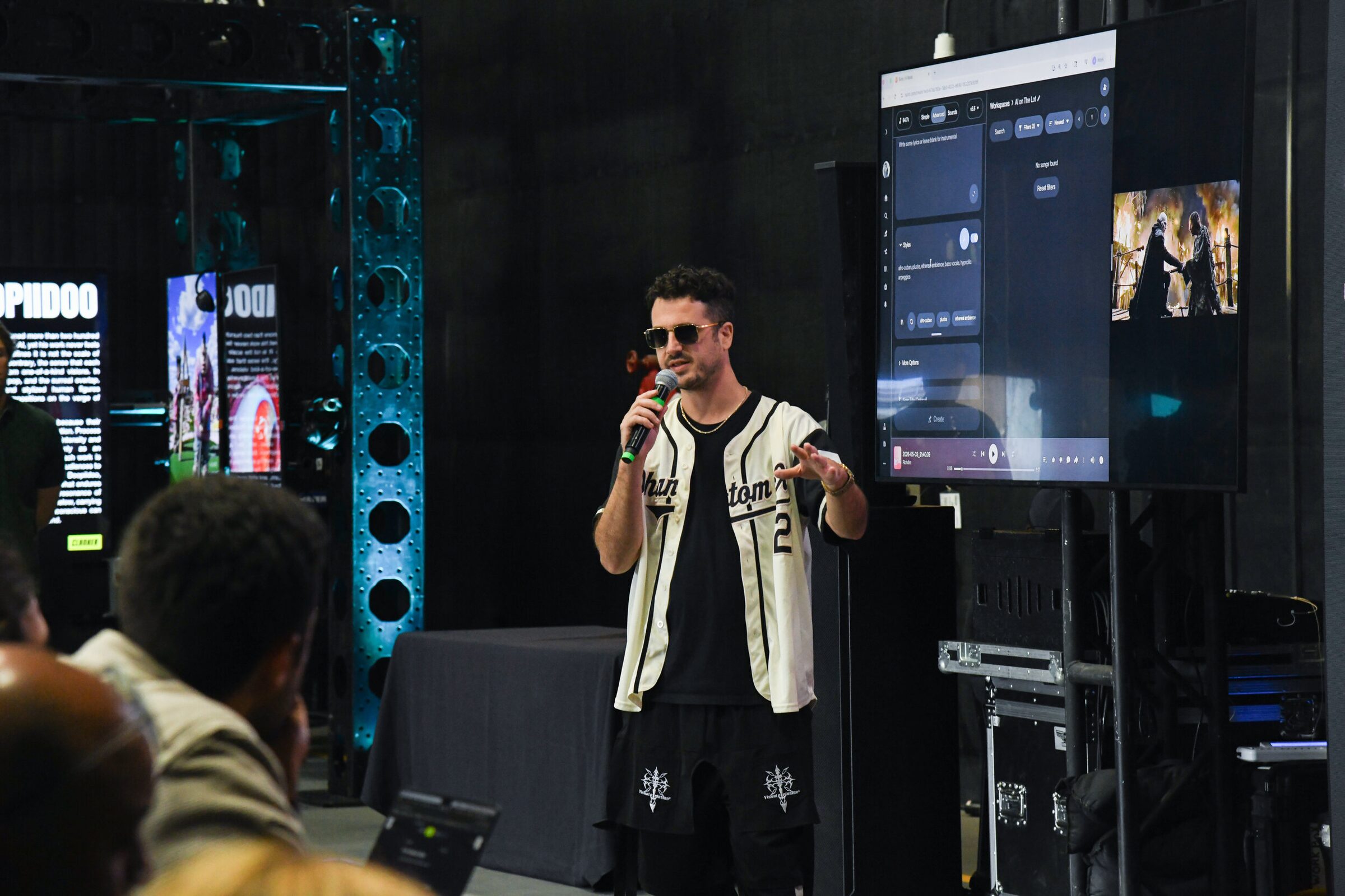 Kavan 'the Kid' Cardoza, in sunglasses and a baseball jersey, talks into a mic beside a screen showing Suno.