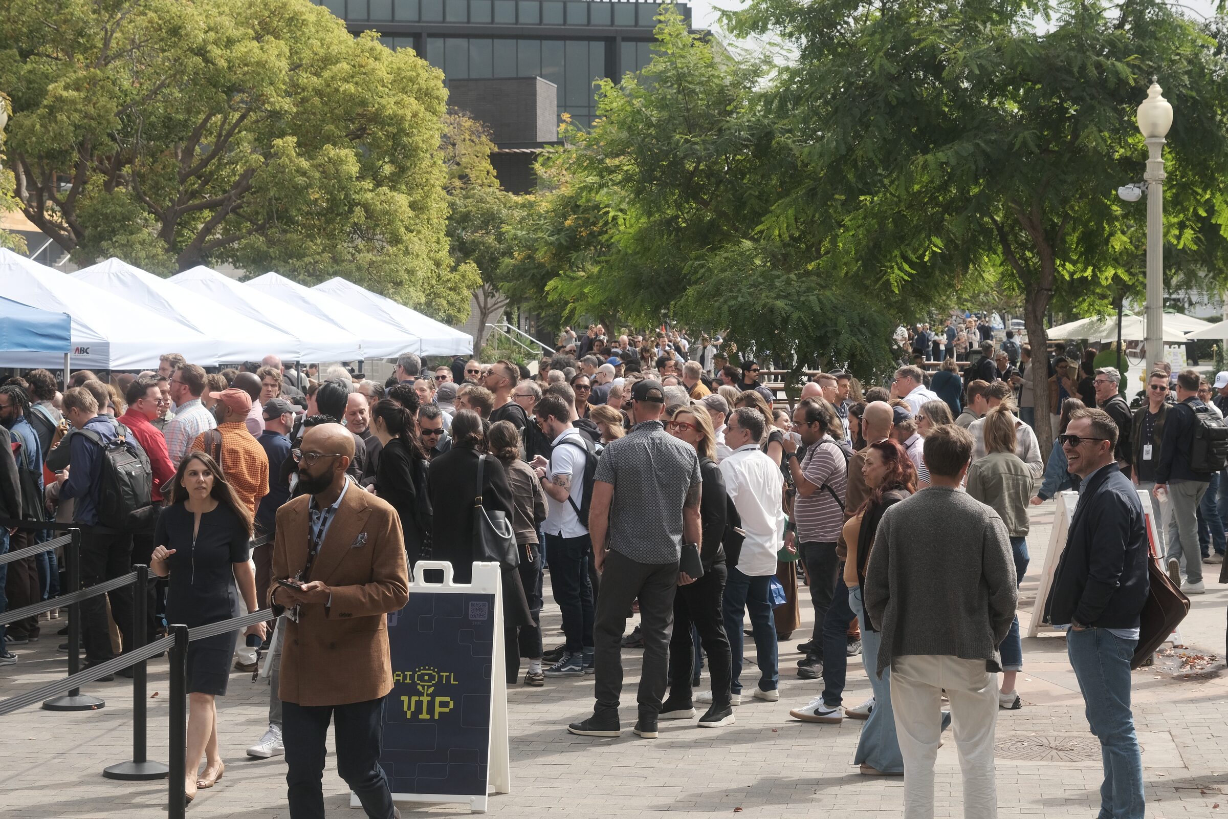 A dense crowd gathers outside a row of white registration tents and a VIP sign under leafy plaza trees.