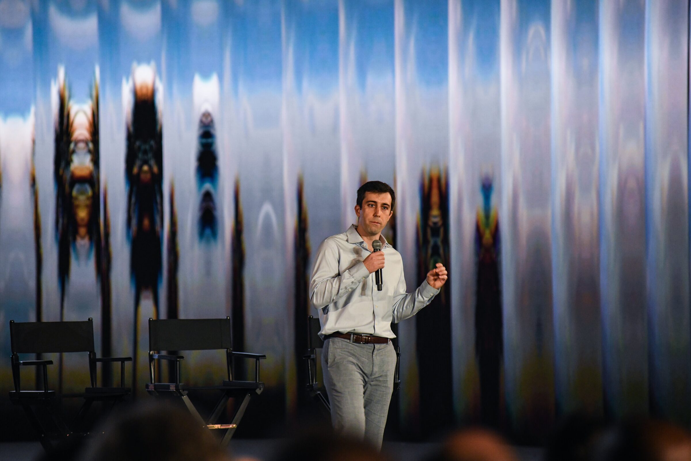 Mike Gioia gestures as he speaks, empty director's chairs beside him on the shimmering projected backdrop.
