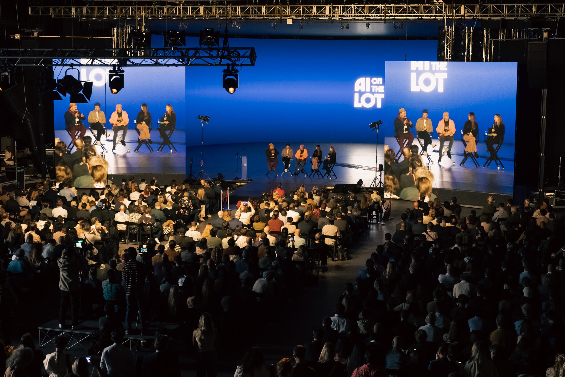 Seen from high up, a full Stage 15 watches five panelists on the blue main stage, shown on two giant screens.