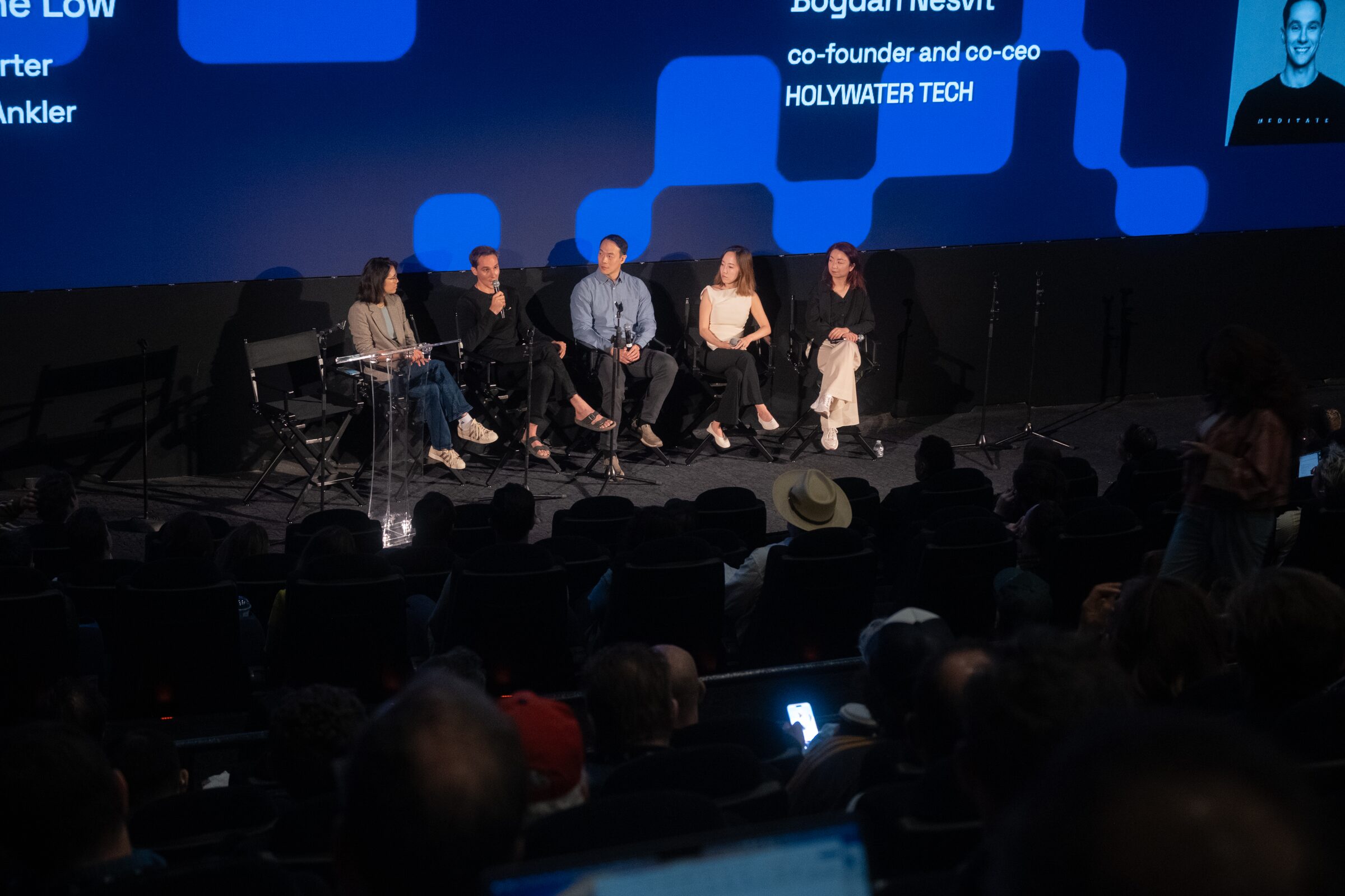 Five panelists in a row on a dark theater stage under a blue screen naming them, the audience in shadow.