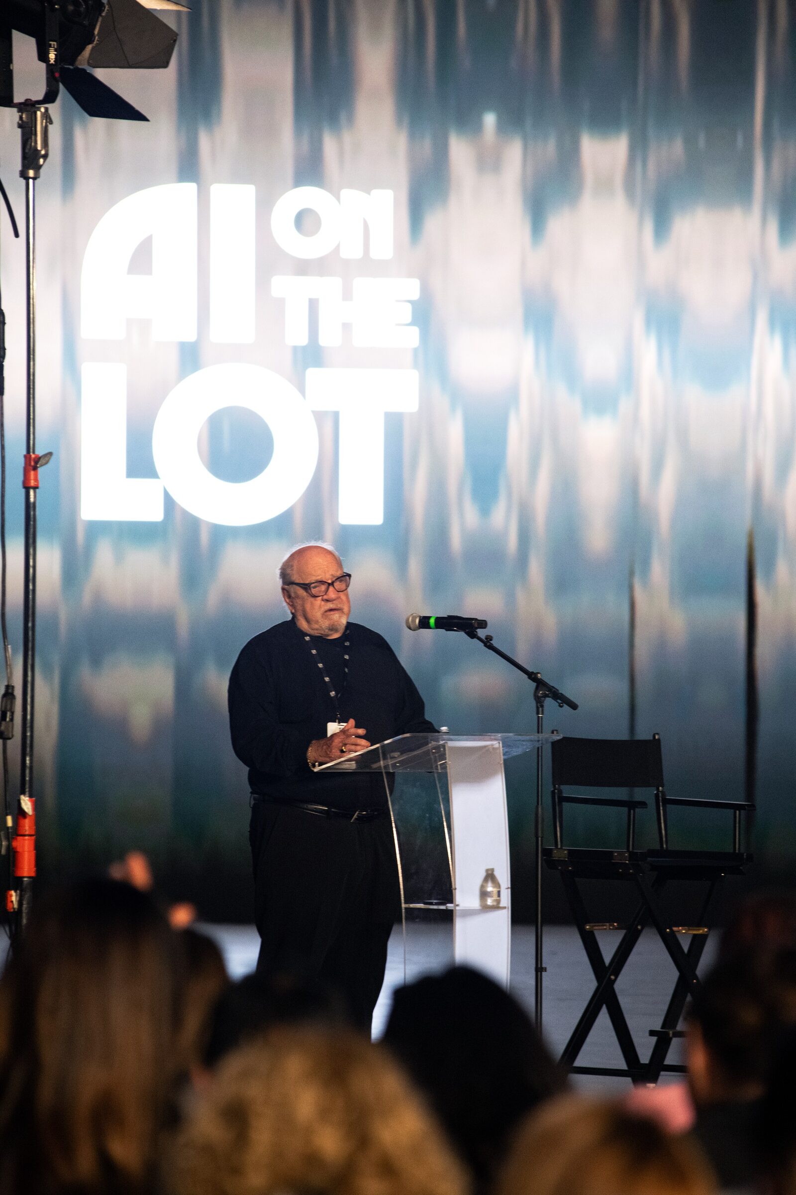 Paul Schrader, in glasses and black, speaks at a clear lectern under the AI on the Lot logo, crowd in front.