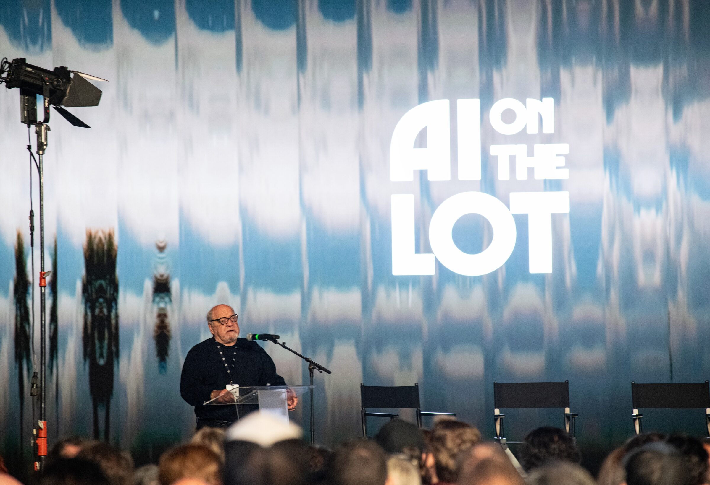 Paul Schrader at a clear lectern below the giant AI on the Lot logo on a shimmering blue projected wall.
