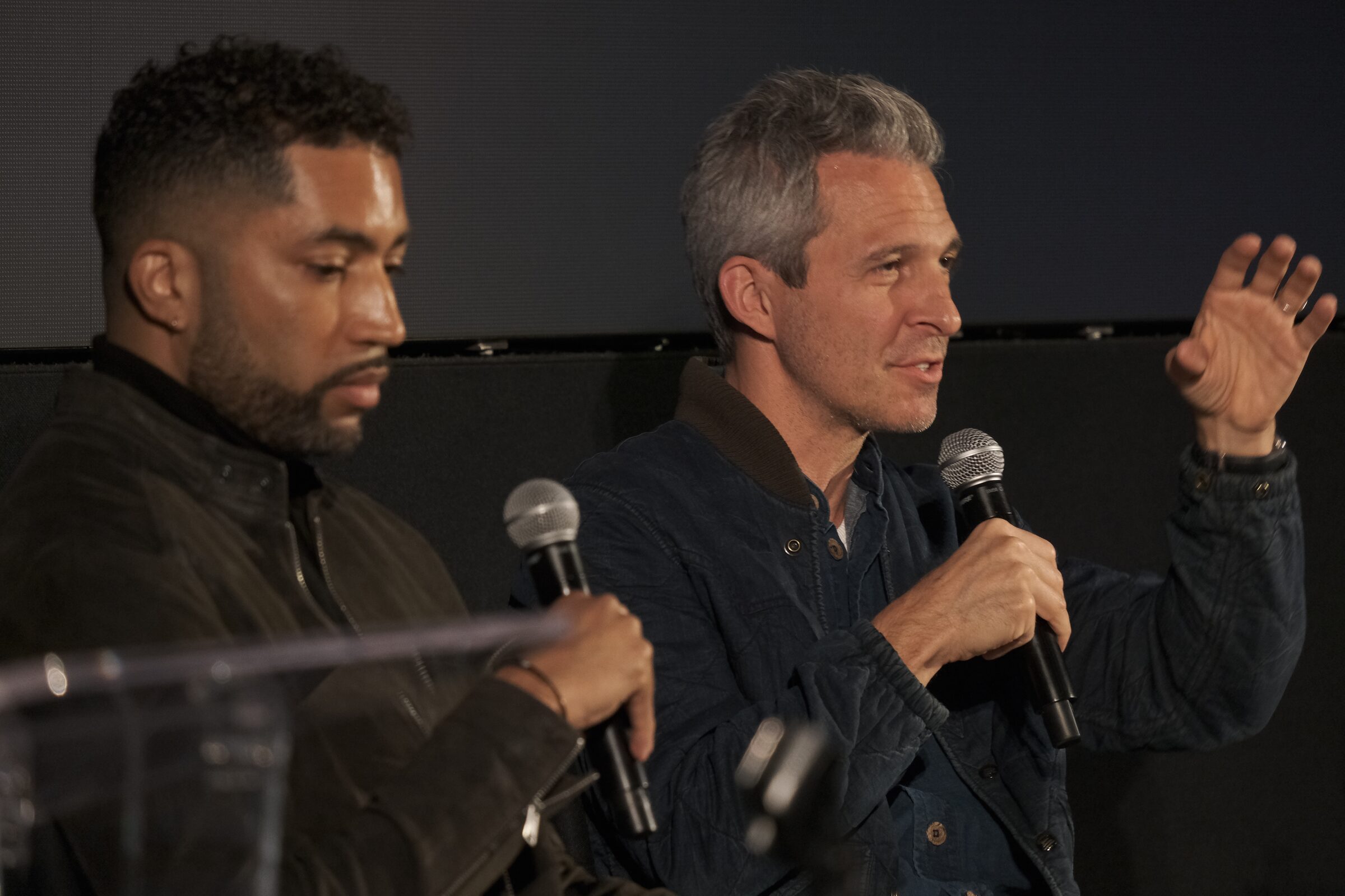Justin Wilkes gestures as he speaks into a mic, a fellow panelist beside him looking down at his own.