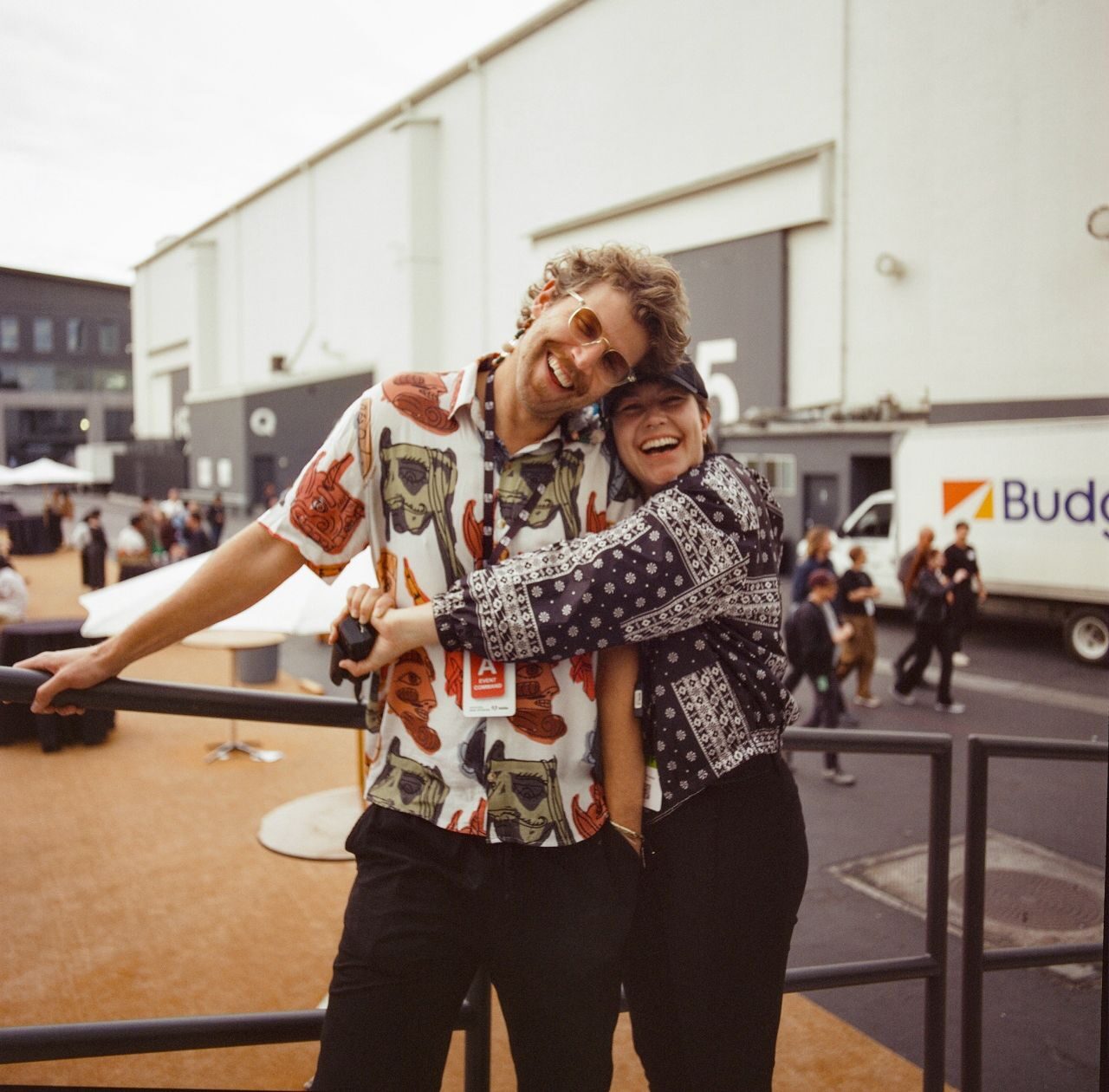 Ames O'Connor hugs a laughing Ian Eck from behind outside a white soundstage, both in patterned shirts.