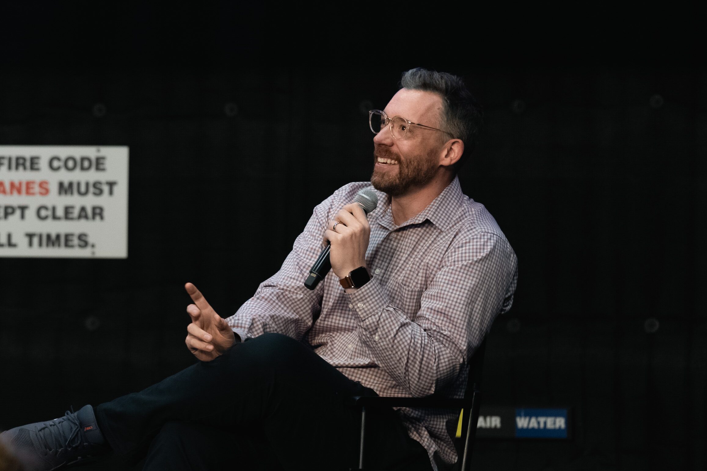 Ted Tremper laughs into a mic in a checked shirt, leaning back in his chair on a dark stage.