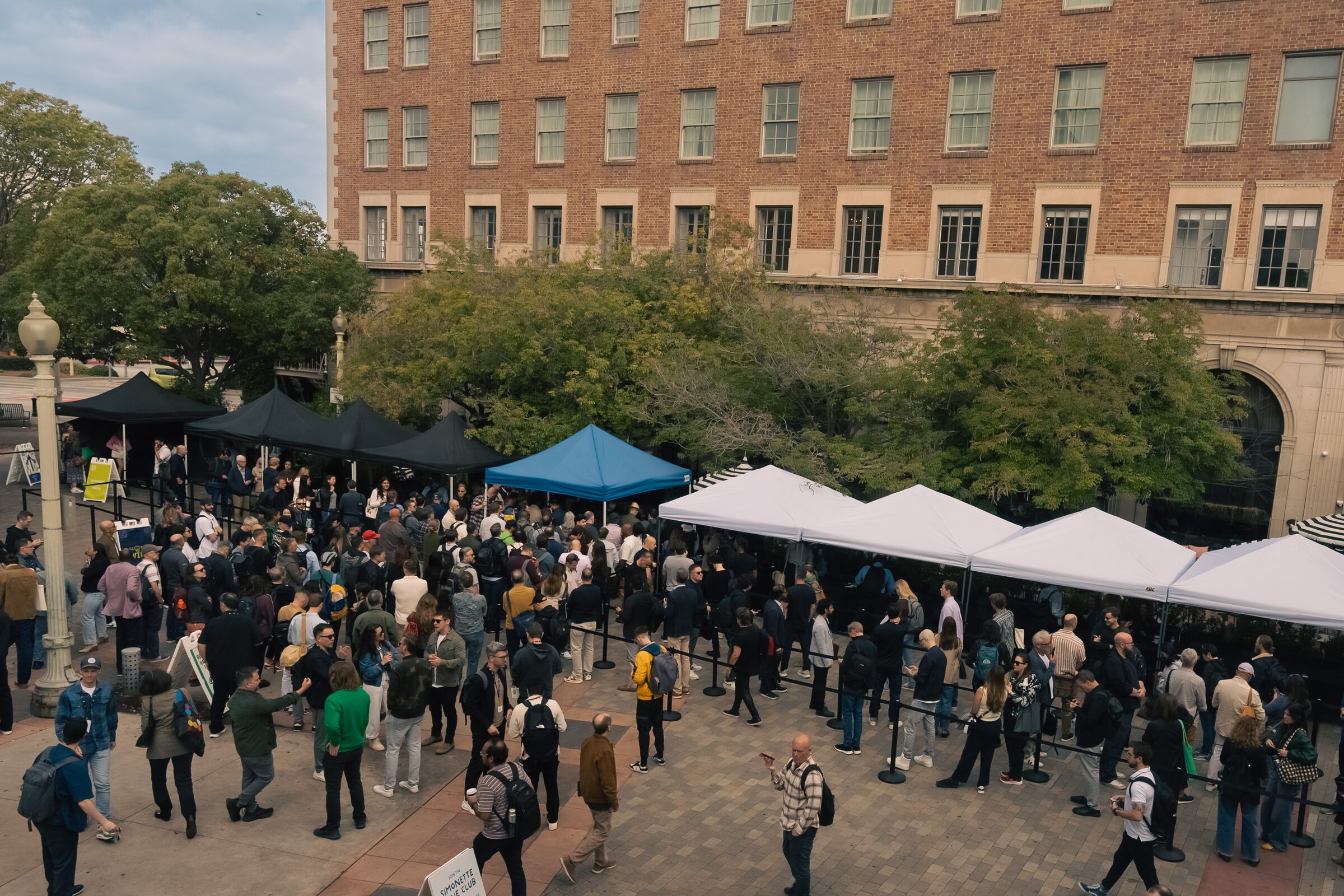 Seen from above, a crowd gathers at white and blue registration tents in front of the brick Culver Hotel.