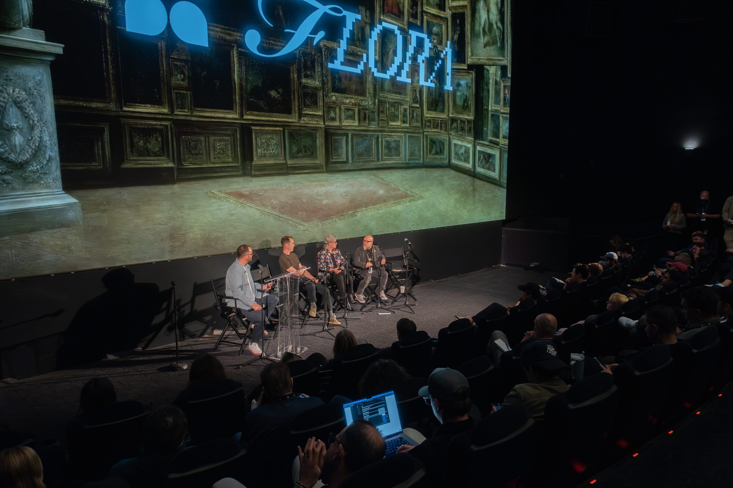 Panelists sit beneath a huge screen of a painted gallery hall with the word "Flora," the theater audience below.
