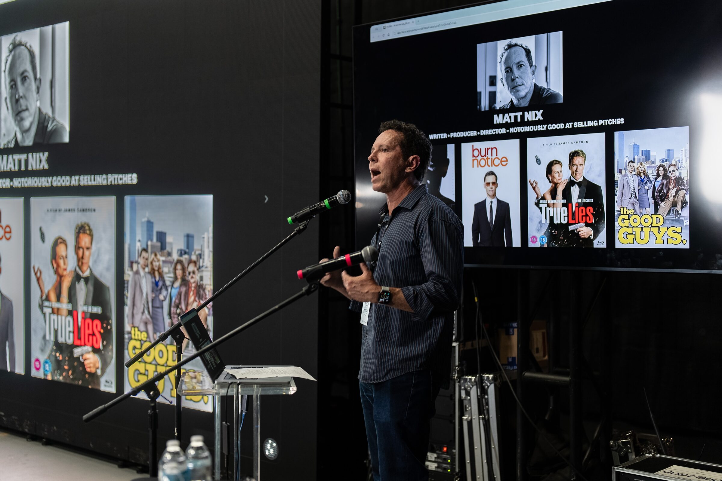 Matt Nix speaks at a mic in front of screens showing his photo and posters for Burn Notice, True Lies and The Good Guys.
