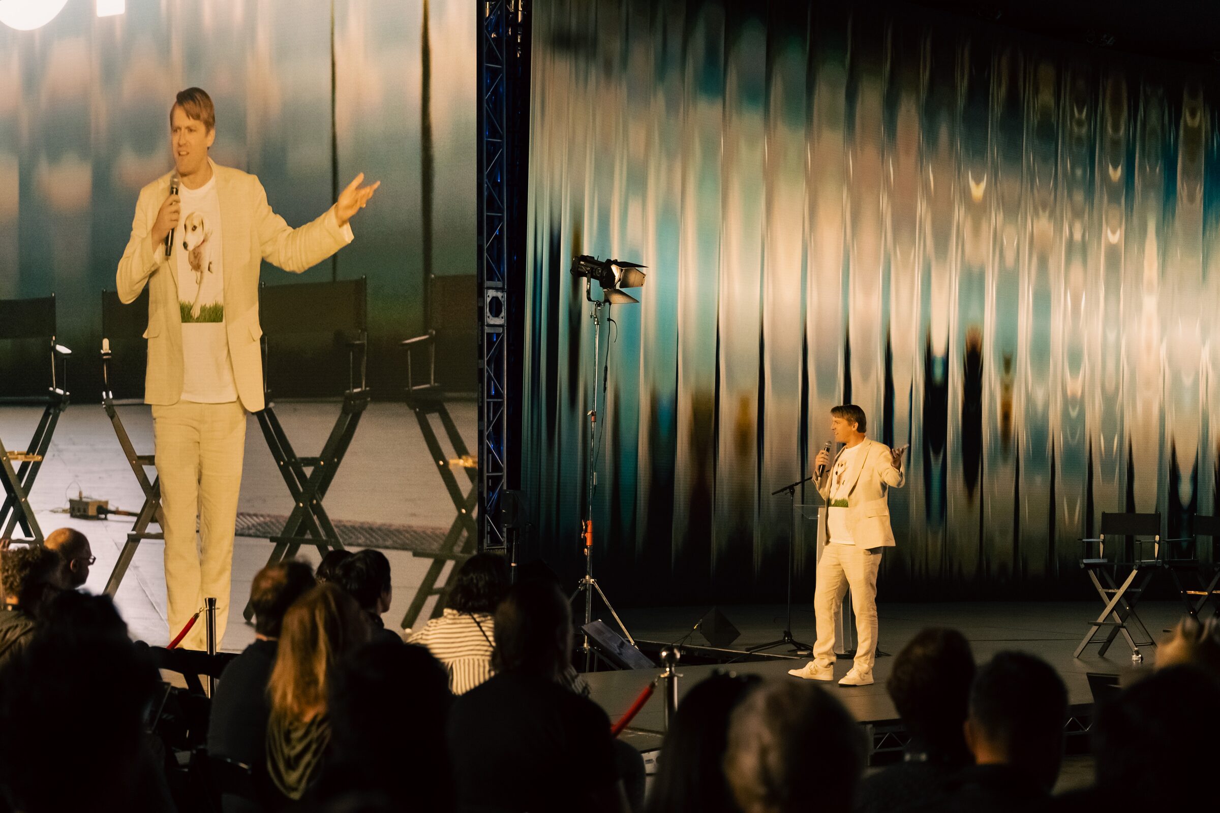 Todd Terrazas, in a cream suit, gestures with a mic on stage and on the giant screen beside him.