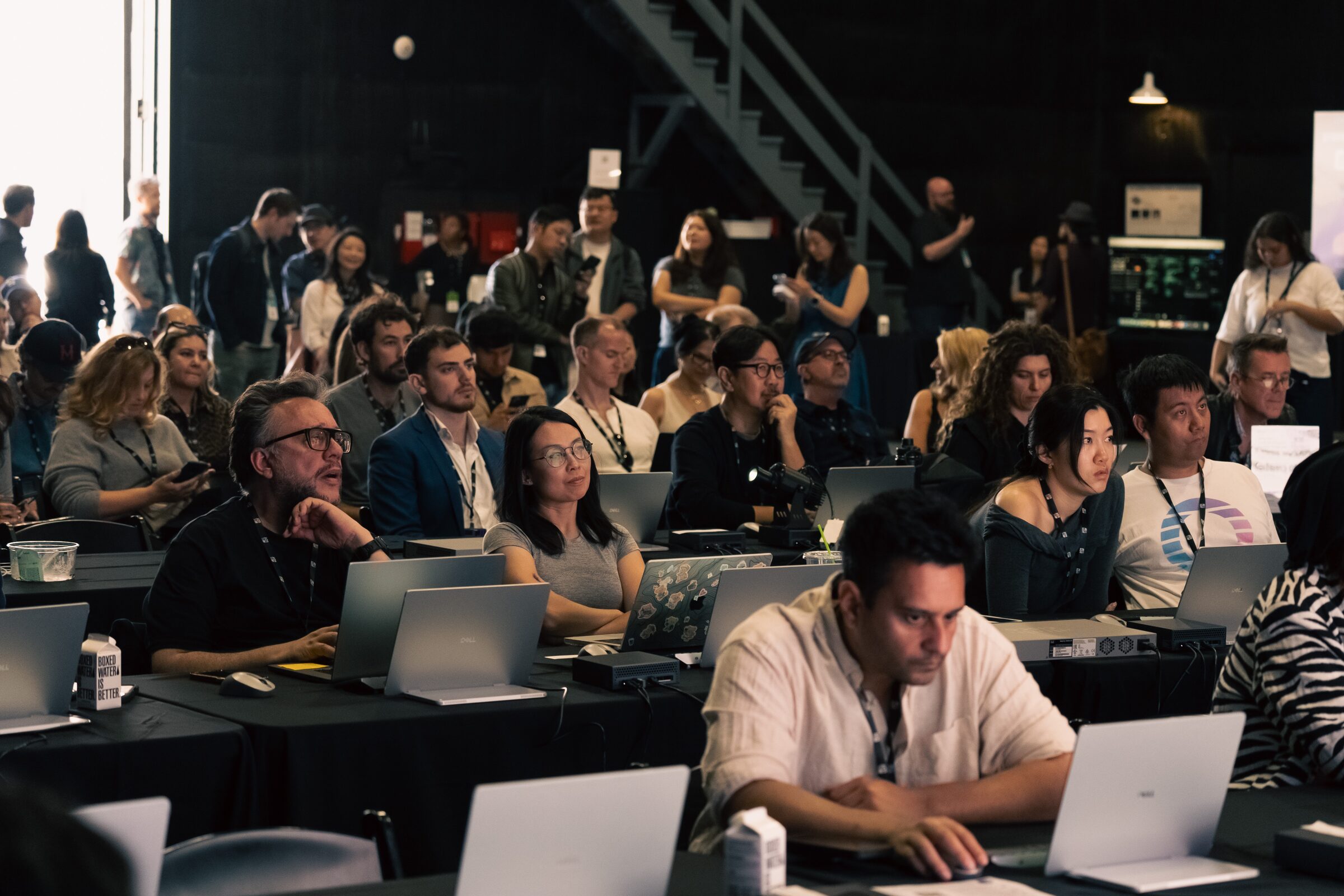 Rows of attendees at long tables work on matching laptops, one in front typing intently, in a dark stage.